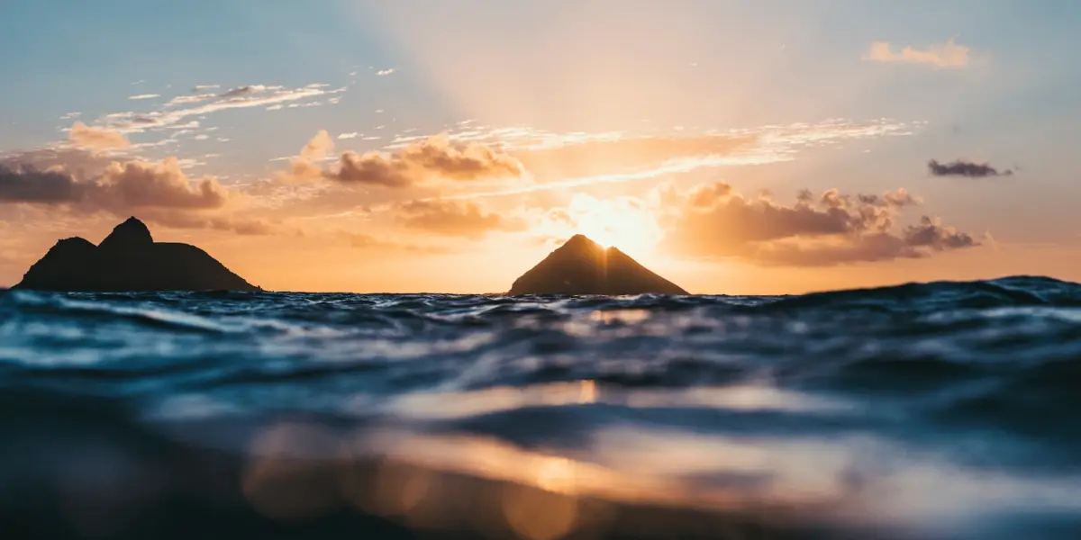 A serene ocean scene at sunset with two small, triangular islands in the middle distance. The sun sets between the islands, casting a golden glow across the sky and reflecting on the water's surface, creating a peaceful and picturesque atmosphere perfect for contemplating your next scuba diving adventure in Oahu.