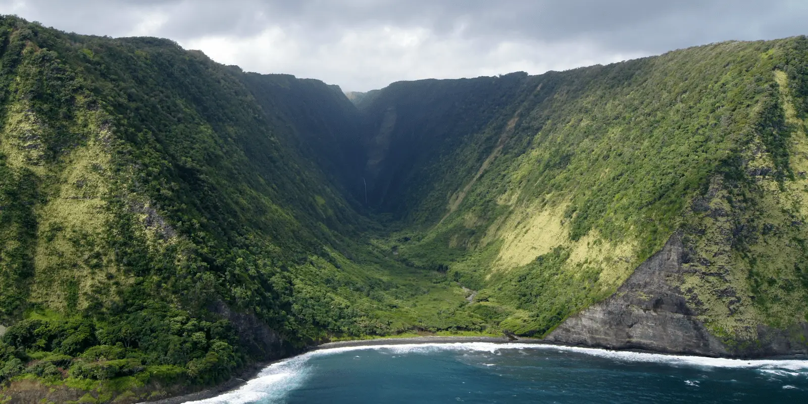 Aerial view of a lush, green valley flanked by steep, forested cliffs descending into the ocean. The coastline features a small, rocky beach with waves crashing onto the shore. The sky above is overcast, casting a soft light over one of the best places for snorkeling on the Big Island.