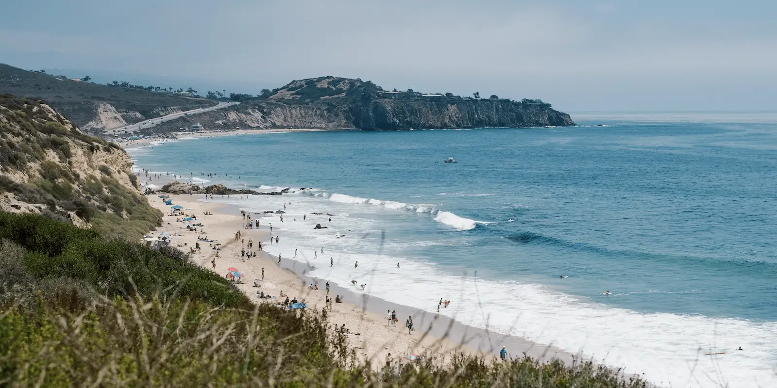 A scenic beach in southern California with white waves crashing against the shore and cliffs in the background. People are scattered along the sandy beach and swimming in the ocean. There are umbrellas and beach towels on the sand. The sky is partly cloudy—perfect timing for some of the Best Snorkeling Southern California offers.