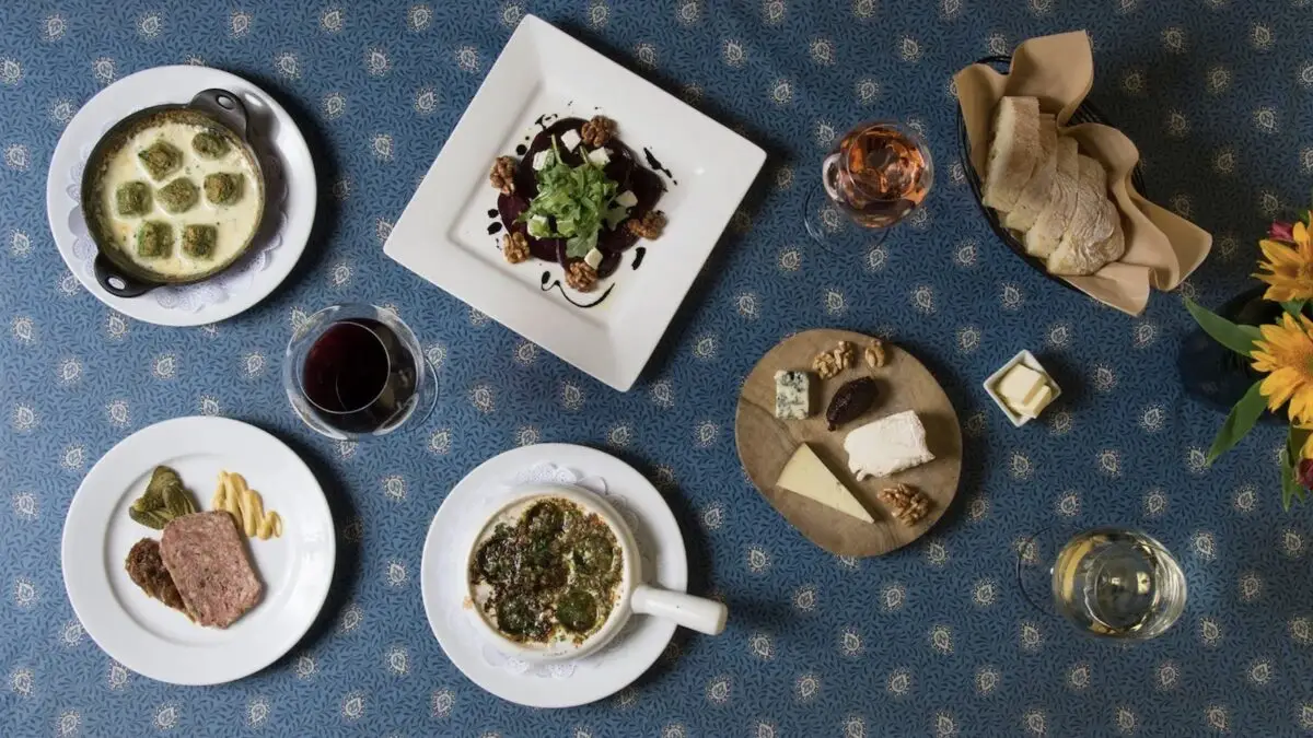 A table set for the best dinner on the Monterey Peninsula features various dishes including soup, bread, cheese, a plate of meat and vegetables, and a dish topped with greens. There are also two glasses of wine and a glass of water. A small vase of flowers is visible in the corner.