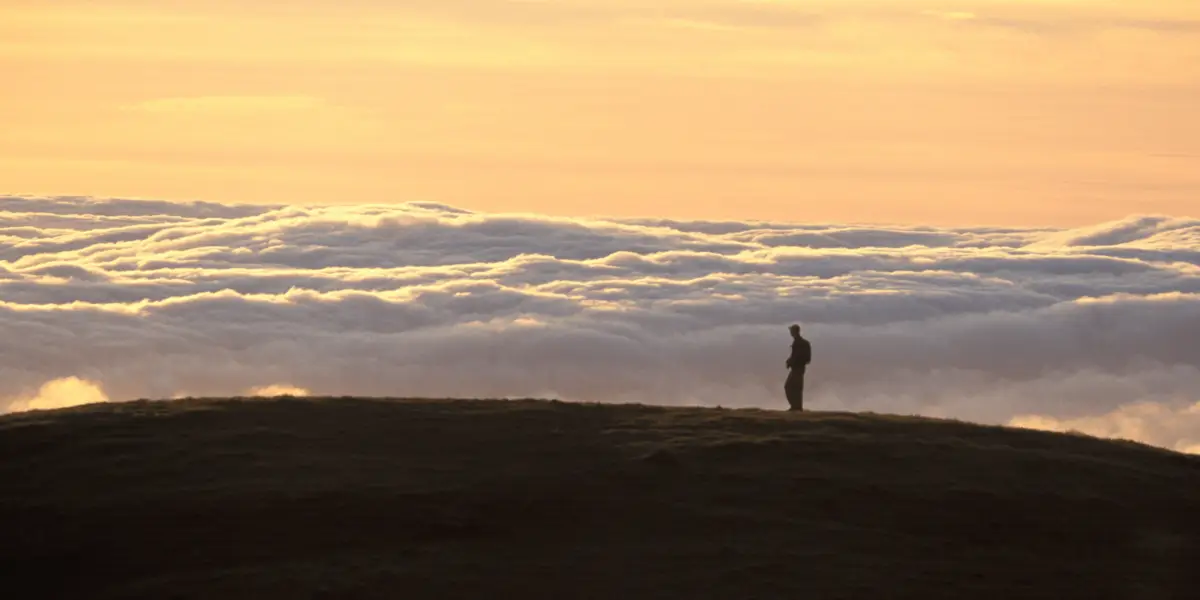 Silhouetted figure standing on a hilltop overlooking a vast sea of clouds, having completed one of the coolest Bay Area summit hikes. The sky is painted in warm hues of orange and yellow, creating a serene and contemplative atmosphere during sunrise or sunset—one of the must-do things to do in North Bay.