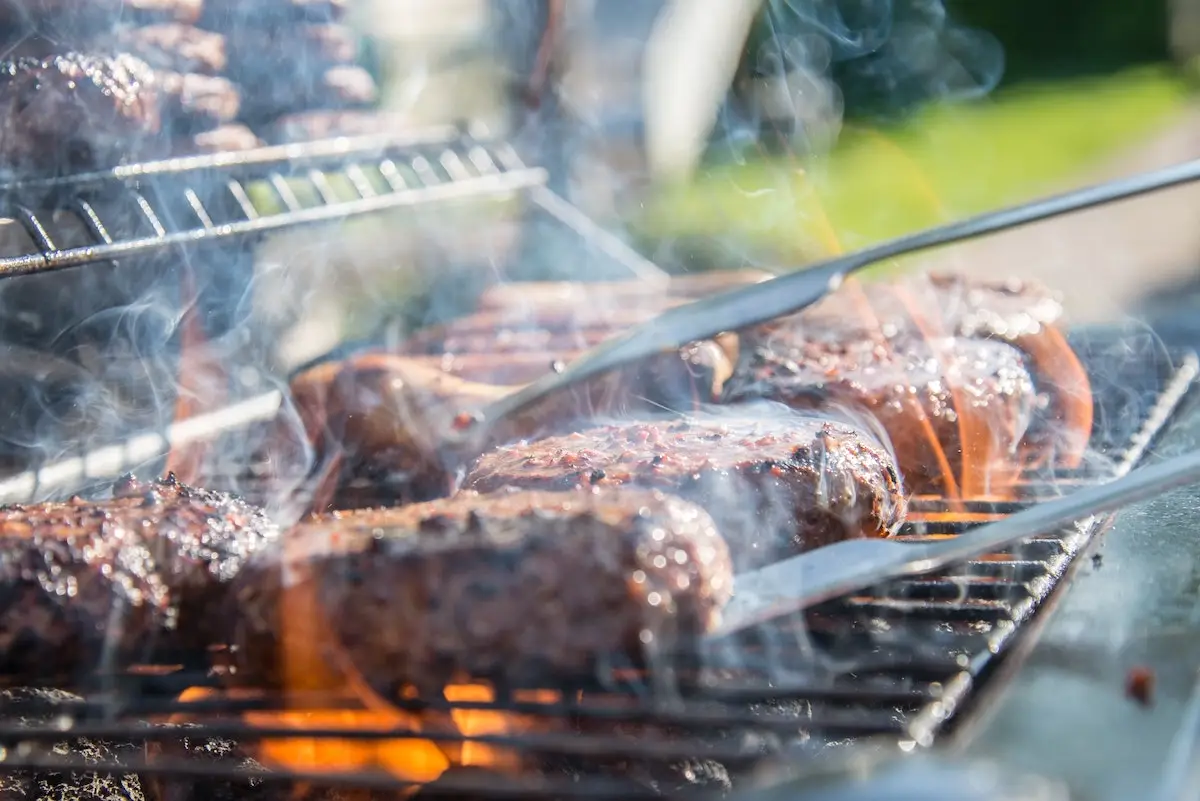 Close-up of a Memorial Day BBQ grill with several pieces of meat being grilled. The meat is sizzling and slightly charred, with smoke and flames visible. Tongs are being used to turn or flip the meat, capturing the festive outdoor cooking atmosphere.