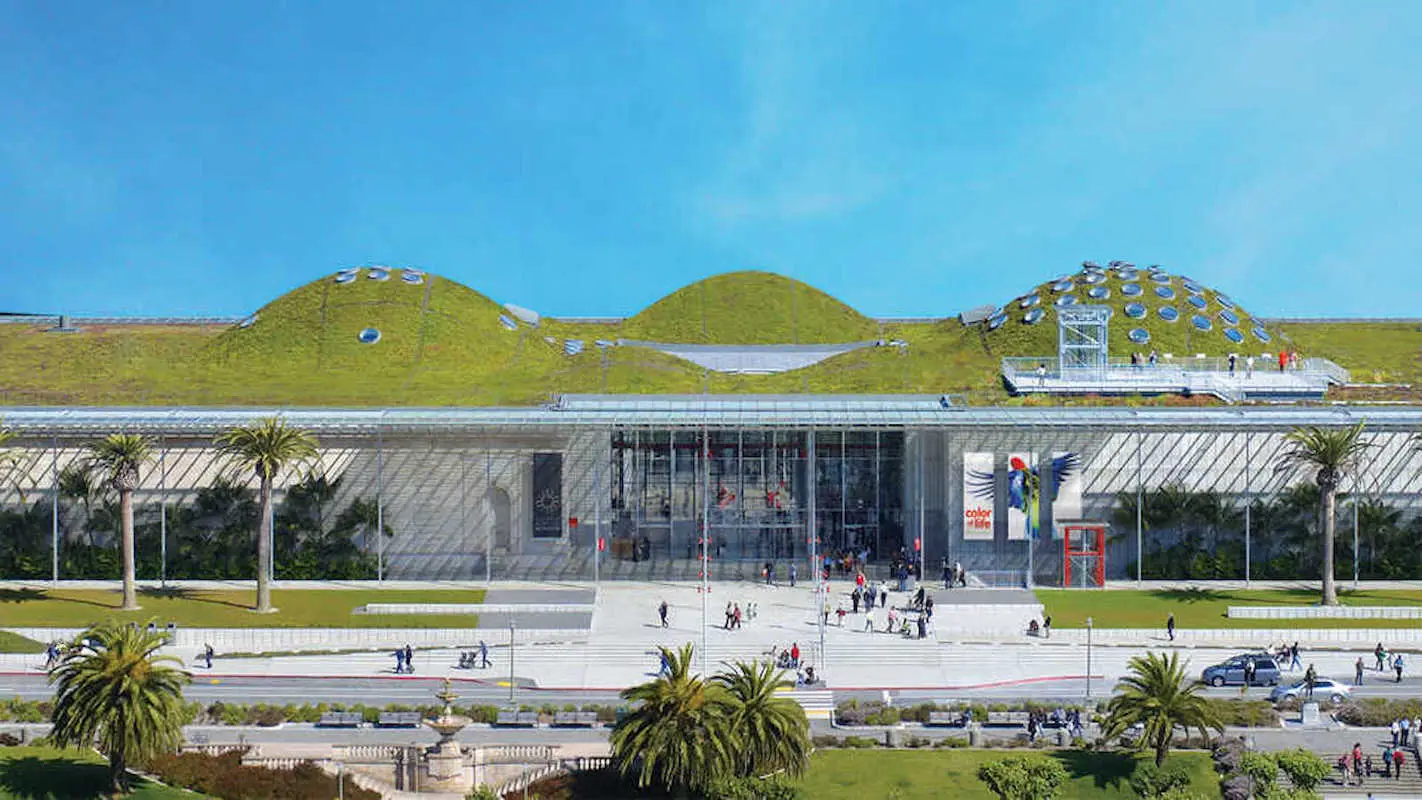 A modern building with a living green roof featuring several rounded domes. The glass facade is surrounded by a plaza where people stroll. Palm trees and green lawns adorn the foreground, with a blue sky overhead, making it one of the coolest kid-friendly museums in Northern California.