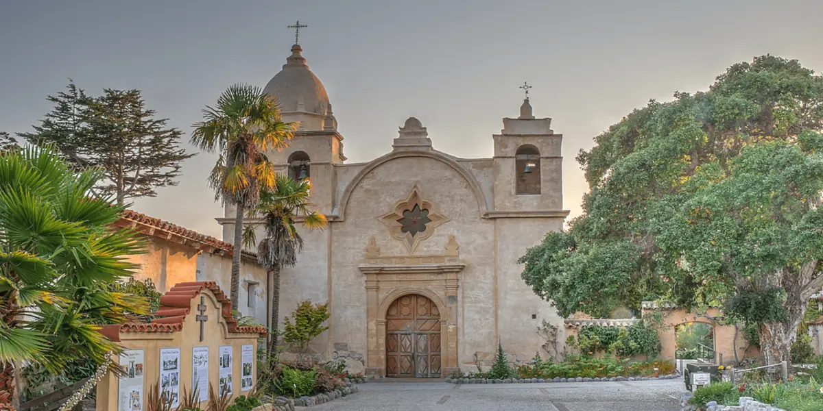 A historic mission-style building reminiscent of the central California missions, featuring a central bell tower and two smaller towers on each side. The facade includes ornate details, a large wooden door, and a cross at the top. Palm trees and lush greenery surround it, with a pathway leading to the entrance.