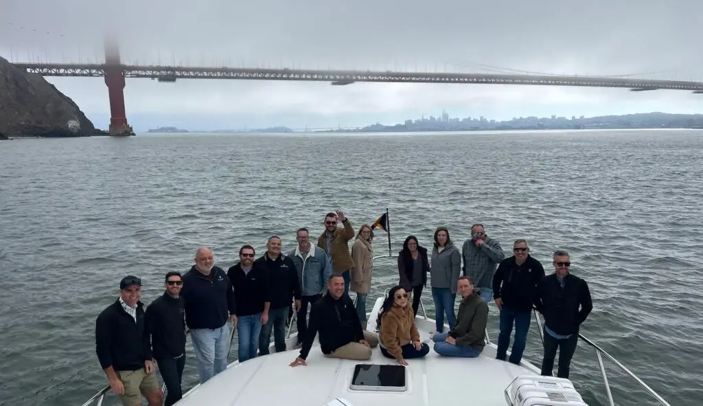 A group of people are gathered on the deck of a charter boat, posing for a photo. The Golden Gate Bridge is visible in the background, shrouded in mist. The water is calm, and the sky is overcast. The group appears to be enjoying a scenic boat trip, ready to live it up.