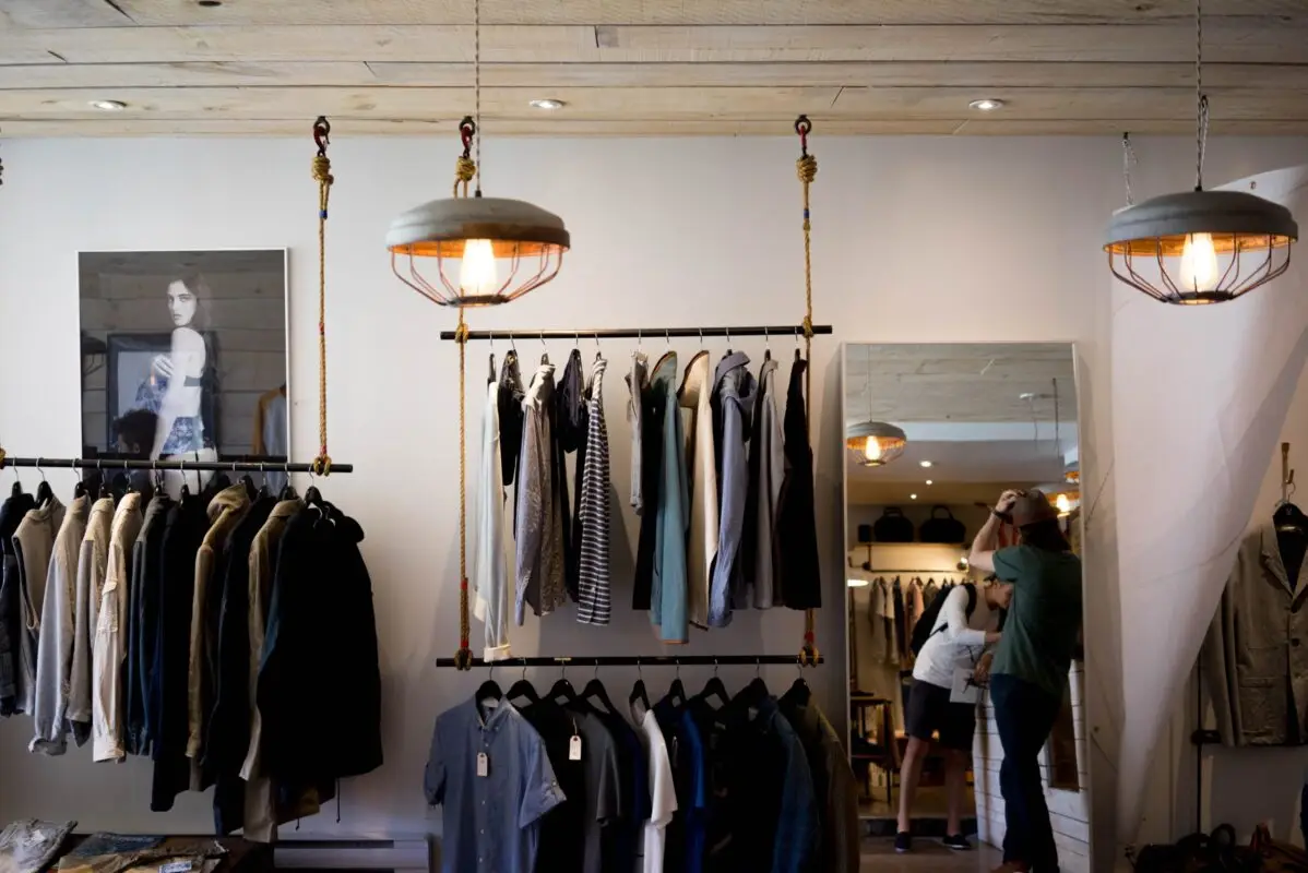 A modern clothing boutique with shirts, jackets, and UV tops for men hanging on racks. Two pendant lights hang from the ceiling. A couple stands near a mirror in the background. A black-and-white photo of a person is displayed on the wall.