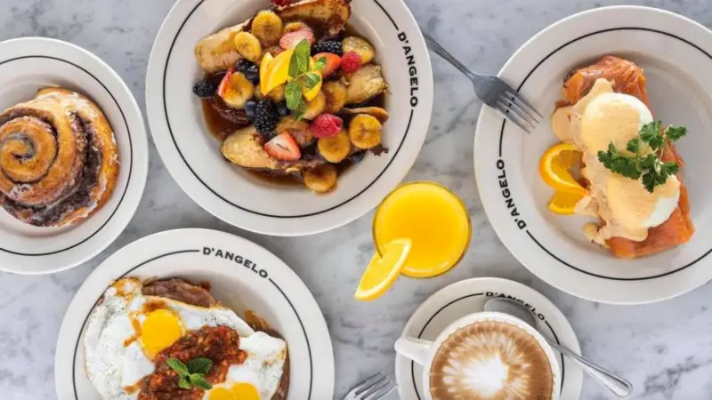 A table set with a variety of breakfast items, showcasing the Best Breakfast in Santa Barbara. Featured dishes include pancakes topped with sliced bananas and berries, eggs benedict on smoked salmon, a cinnamon roll, and a plate of eggs over chilaquiles. Also on the table are a glass of orange juice and a cappuccino.