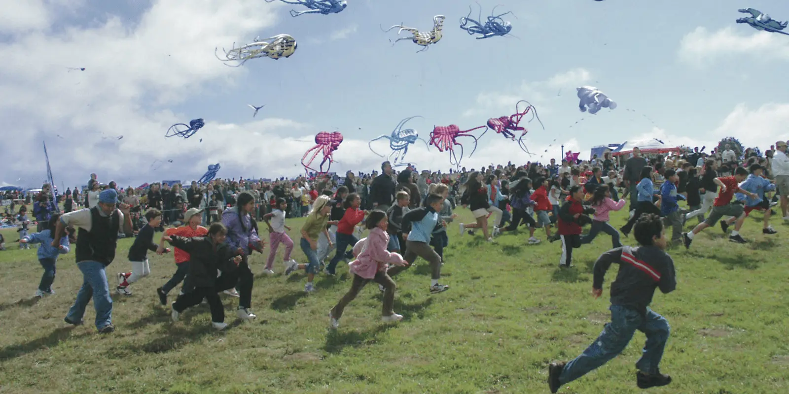 A large crowd of people, including many children, run and play on a grassy field. High above them, colorful kites in various shapes and sizes are flying against a partly cloudy sky. The scene is lively and joyous, capturing one of the Best Annual Events in the East Bay with many participants engaged in the festivities.