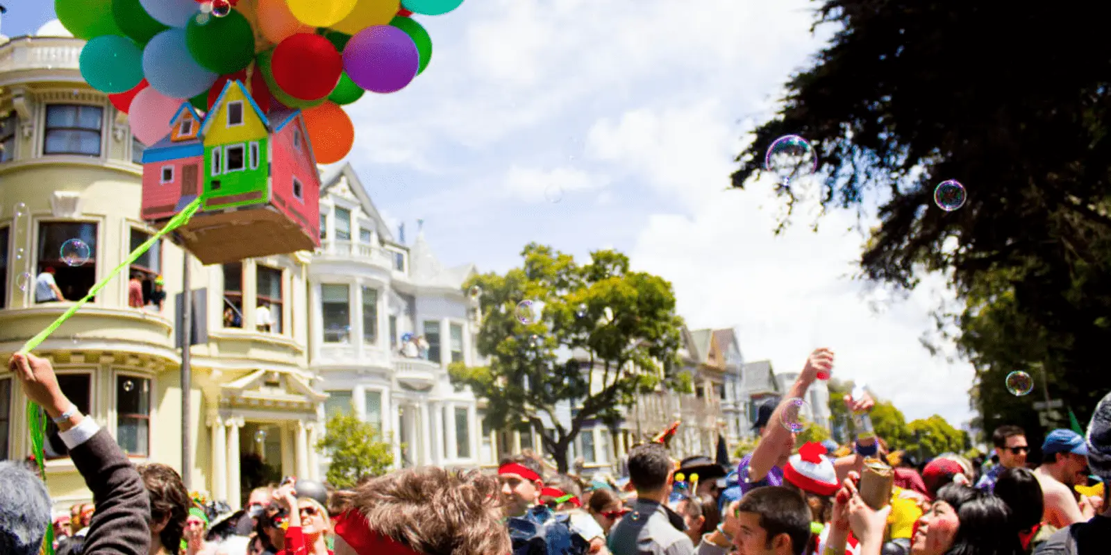 A colorful parade scene with people cheering and blowing bubbles. A float designed as a house from the animated movie "Up" is attached to a bunch of multicolored balloons, floating above the crowd. Victorian-style houses and trees are visible in the background, making it one of the Best Annual Events in San Francisco.