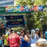 A lively crowd of people dances energetically in front of a stage at an outdoor summer festival. The stage banner reads "Salsa Stage" at San Jose Jazz Summer Fest, one of the Best Annual Events in the South Bay. Trees and modern buildings can be seen in the background under a clear, sunny sky.