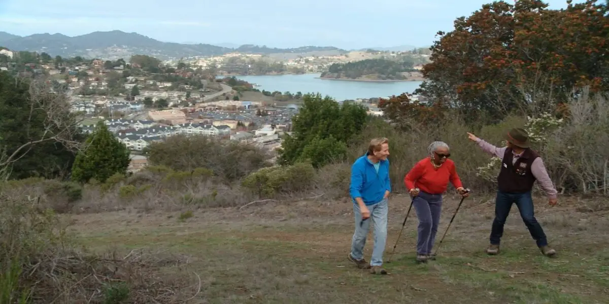 Three people are hiking on a trail overlooking a scenic view of a coastal town and hills. Doug McConnell from "Bay Area Backroads," the older man, and the younger man use walking sticks, while the third person gestures toward the vista. Trees and shrubs line the hillside pathway.