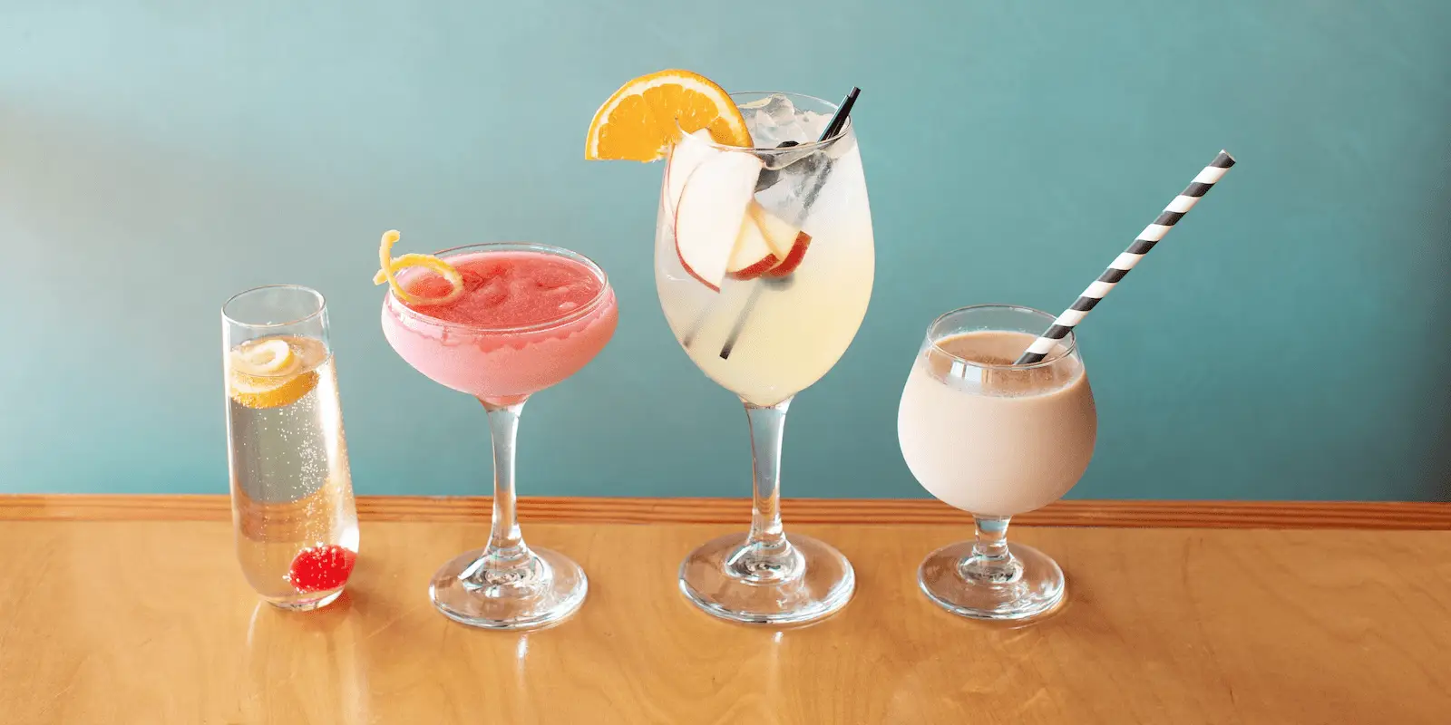 A row of four different drinks on a wooden surface against a light blue background, perfect for East Bay Happy Hour. From left to right: A flute glass with a clear bubbly drink and lemon twist, a martini glass with a pink frozen drink and lemon twist, a wine glass with a yellow drink, ice, and orange slice, and a round glass with a creamy drink and striped straw.