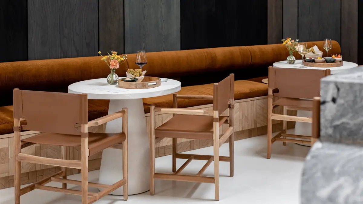 A modern café inspired by Sonoma Tasting Rooms features minimalist décor with sleek marble tables, wooden chairs with brown leather cushions, and a long brown cushioned bench. Tables are adorned with flowers, glasses of red wine, small snacks, and neatly arranged utensils.
