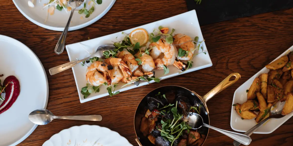 A table setting featuring a white rectangular plate with cooked shrimp garnished with lemon slices and greens. Surrounding it, there are other dishes, including roasted vegetables in a black bowl and golden-brown potatoes in a white bowl - perfect for when Bay Area chefs eat out.