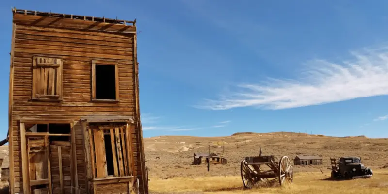 A weathered, tilted wooden building stands in an arid landscape under a clear blue sky, reminiscent of Bodie, California Ghost Town. An old wooden cart sits nearby. In the background, more dilapidated wooden structures are scattered across the dry terrain.