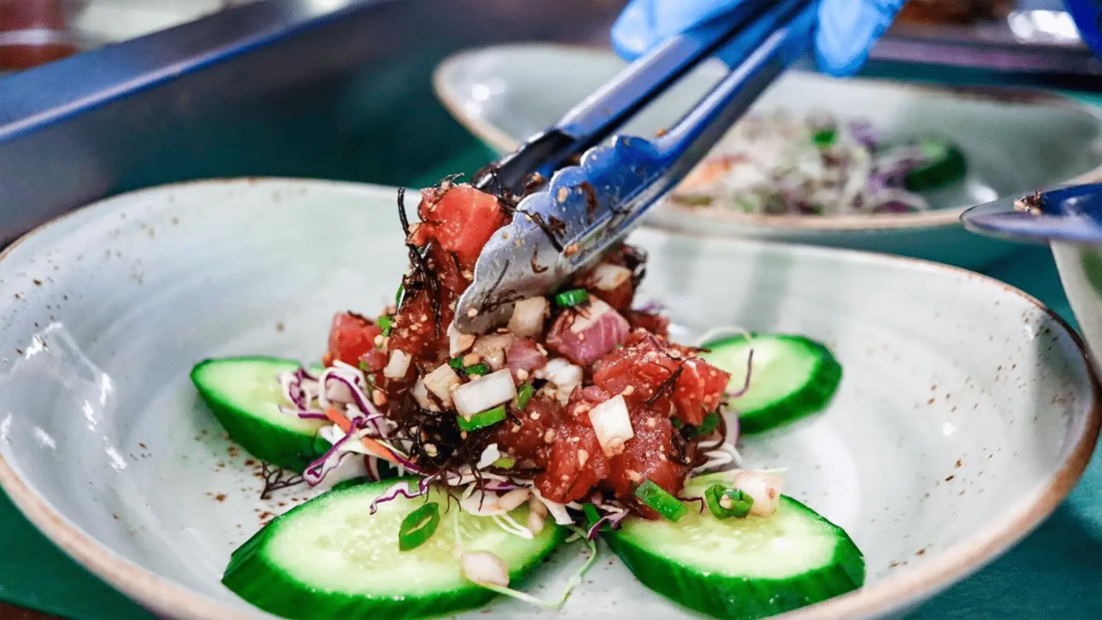 A person using tongs to place a portion of Best Poke Maui on a plate. The dish includes diced tuna, chopped onions, green onions, and sesame seeds, arranged atop a bed of shredded vegetables and surrounded by cucumber slices. The person is wearing blue gloves.
