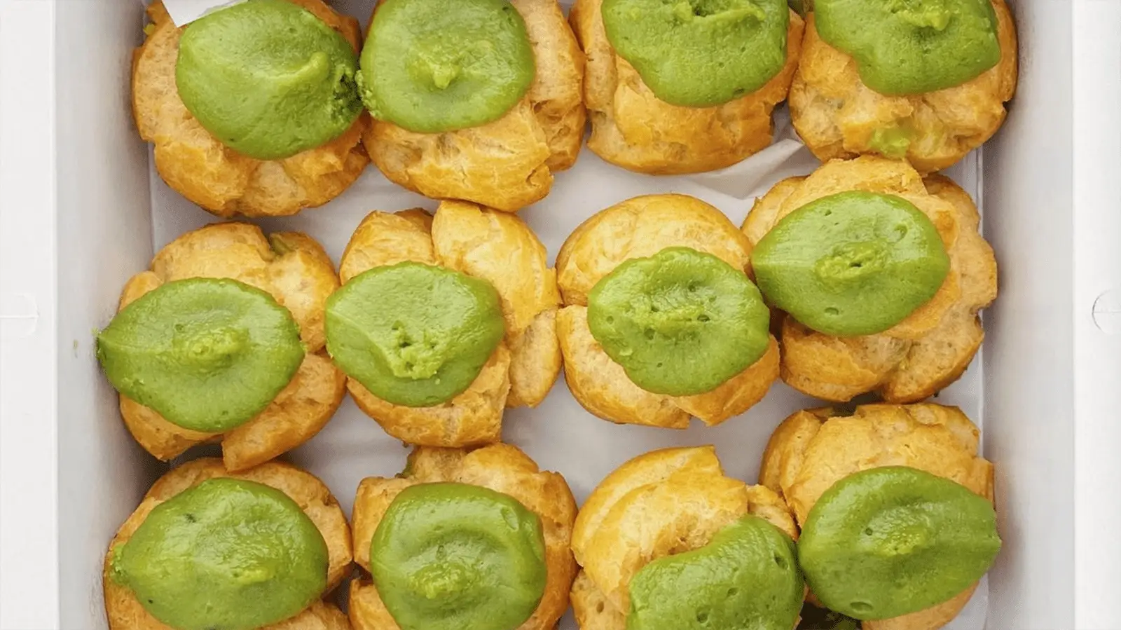 A white rectangular box containing twelve cream puffs, each golden-brown and topped with green matcha cream, creating a colorful and appetizing display. These delectable treats are among the best desserts Oahu has to offer.