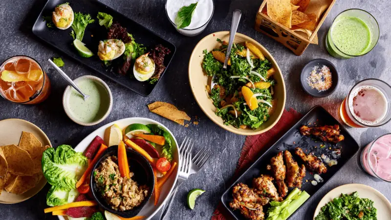 A colorful spread of various dishes on a dark surface, featuring stuffed eggs, a mixed salad with oranges, a bowl with rice and vegetables, green dipping sauce, chicken wings, drinks, tortilla chips, and a bowl of fresh greens—the best dinner Ko Olina has to offer.