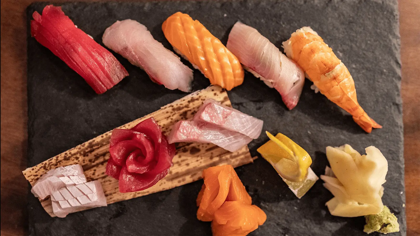 A slate serving board with assorted sushi pieces is displayed, showcasing the best Sushi central Maui has to offer. The arrangement includes tuna, salmon, yellowtail, and shrimp nigiri on top, along with various sashimi pieces alongside pickled ginger and wasabi at the bottom. The sushi is vibrant and fresh.