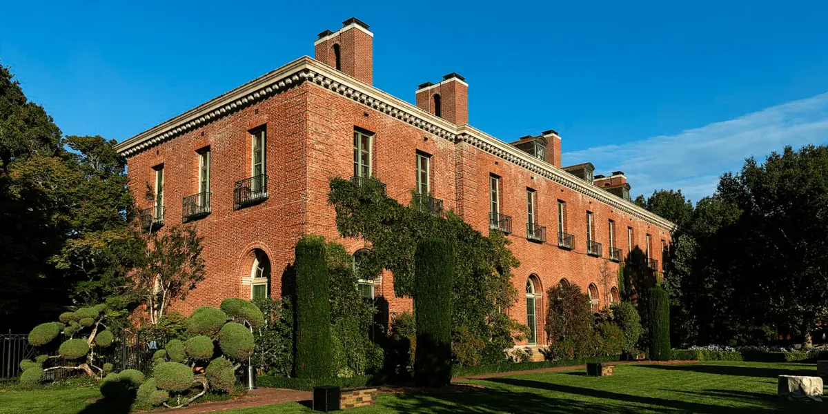 A large brick building with tall, arched windows and several chimneys stands against a clear blue sky. The structure, reminiscent of the Filoli Mansion Gardens, is surrounded by well-maintained shrubbery, topiary, and a lush green lawn. The building's facade features ivy and climbing plants.