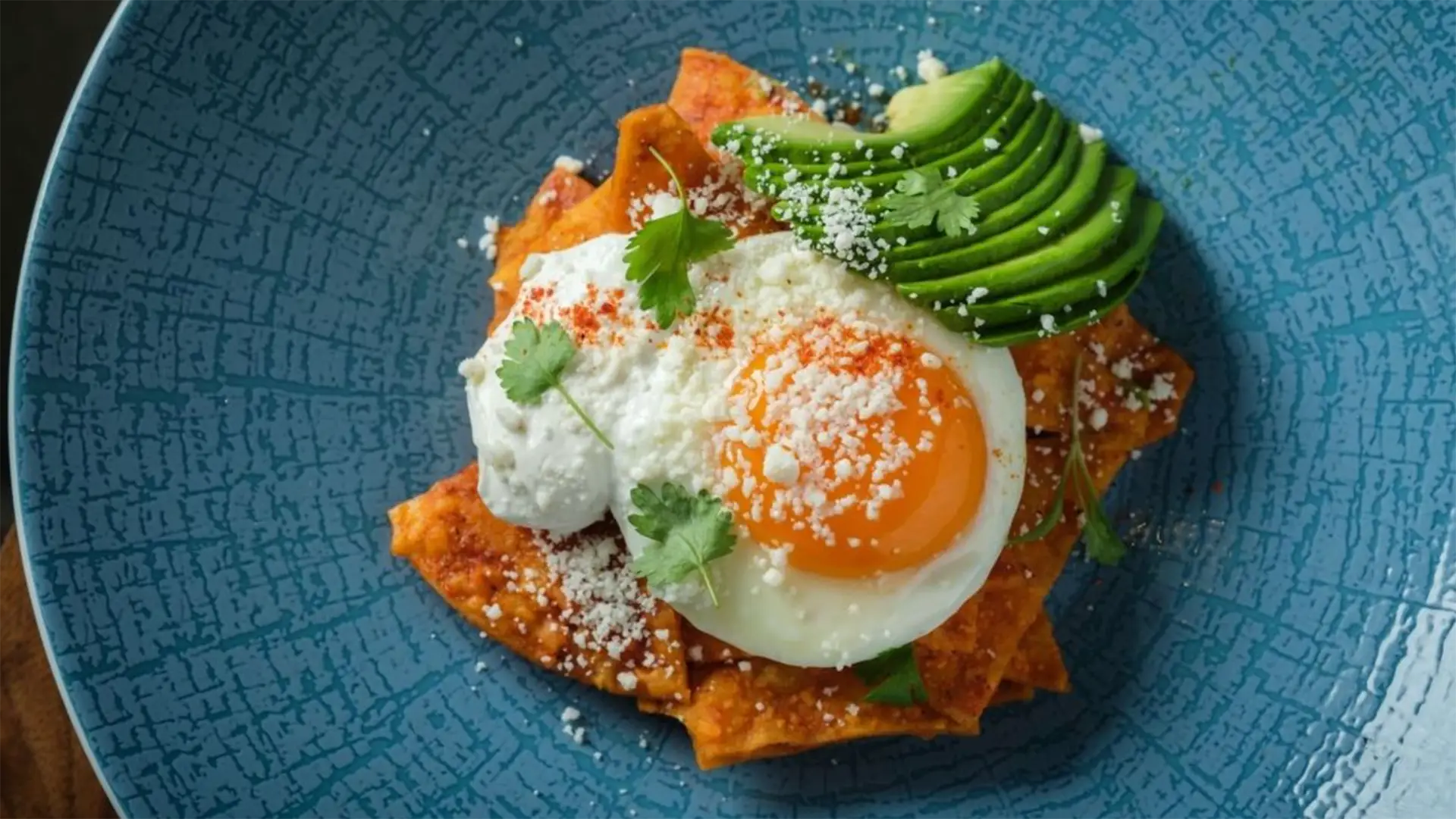 A vibrant dish on a blue plate featuring chilaquiles topped with a sunny-side-up egg, sliced avocado, a dollop of sour cream, crumbled cheese, cilantro leaves, and a dusting of paprika—truly the best breakfast Santa Barbara has to offer.