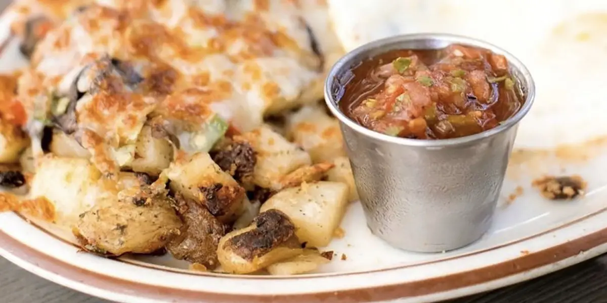 A plate with a serving of roasted potatoes and vegetables topped with melted cheese, next to a small metal cup filled with salsa. The food is on a white dish with a brown rim, making it the best breakfast Tahoe has to offer.