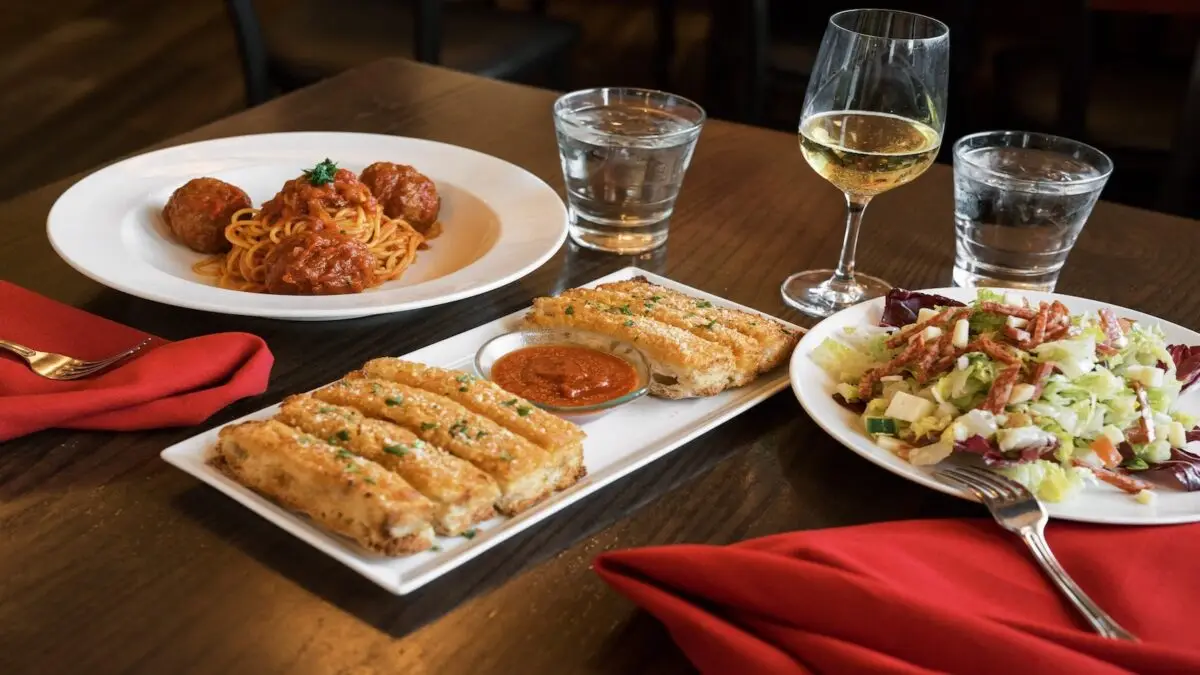 A restaurant table with a plate of spaghetti and meatballs, a glass of white wine, two glasses of water, a white plate of salad with bacon pieces, and a rectangular plate with garlic breadsticks and marinara sauce, all set on a dark wood surface with red napkins—truly the best dinner you’ll find on the Monterey Peninsula.