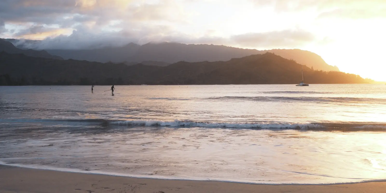 A serene Hanalei beach scene at sunset with gentle waves lapping the shore. Two paddleboarders glide across the calm water—one of the top ways to get in the water—and a sailboat is anchored further out. The sky is a mix of warm sunset hues and clouds, with mountains silhouetted in the background.