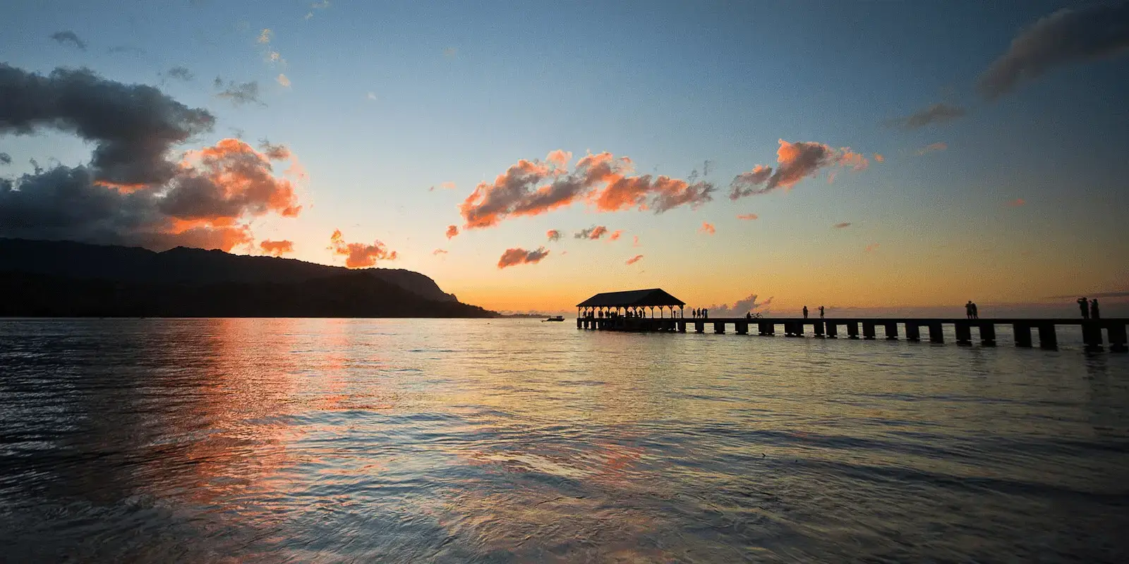 A serene sunset over a tranquil ocean, with a pier extending into the water. The sky is painted in shades of orange and pink, reflecting on the water's surface. Silhouettes of people are visible on the pier, as if they've just returned from one of the top island tours in Kauai. Mountains frame the background.