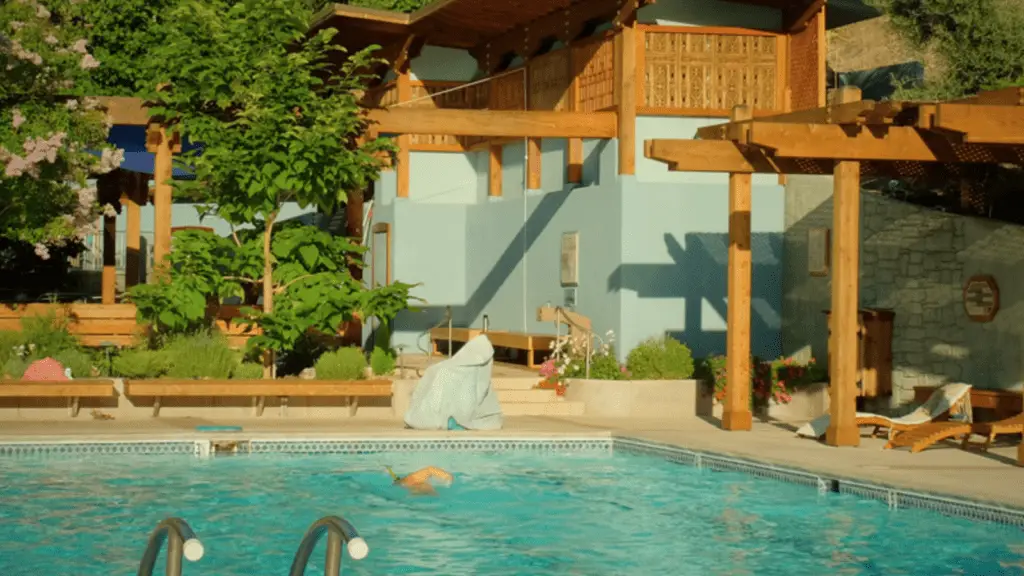 A person is swimming in a clear blue pool surrounded by wooden pergolas and lush greenery. A modern light blue house with wooden accents overlooks the pool. The warm sunlight casts shadows across the scene, creating a serene and inviting atmosphere reminiscent of the Best Spa and Wellness Hotels in the Bay Area.