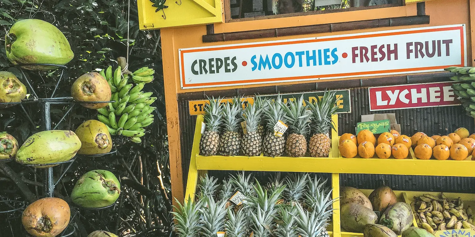 A vibrant fruit stand offers fresh produce, including hanging coconuts, bunches of bananas, pineapples, and lychees. A colorful sign above reads "Crepes, Smoothies, Fresh Fruit." The stand is adorned with various tropical fruits invitingly displayed alongside foods that define Hawaiian culture.