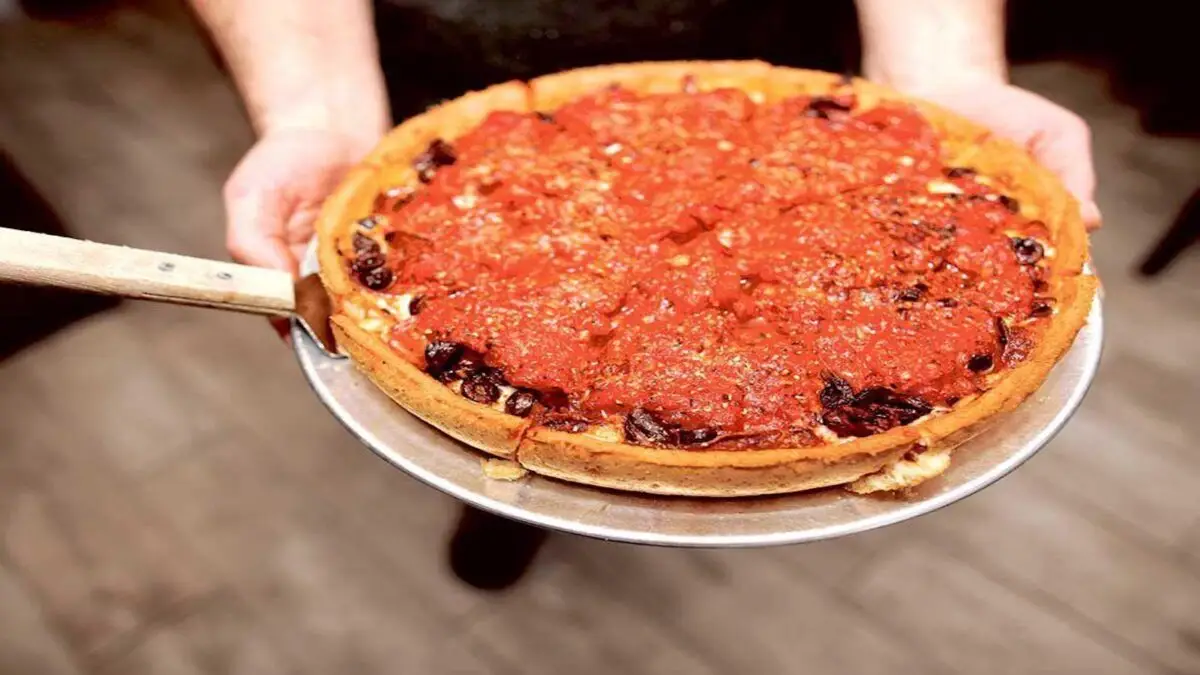 A person holds a freshly made Chicago-style deep-dish pizza on a metal tray. With its thick, golden-brown crust, generous layer of chunky tomato sauce, and visible pieces of sausage and olives, it’s undoubtedly set to be the best dinner on the Monterey Peninsula.