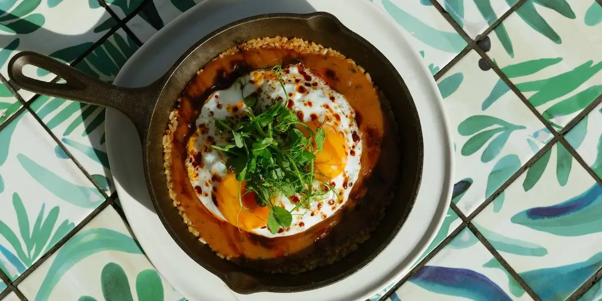 A skillet filled with a dish topped with two fried eggs, spicy red sauce, and fresh green herbs is placed on a white plate. The skillet appears to be on a patterned, green and white tile surface. This vibrant and flavorful dish could easily rival the best loco moco Oahu has to offer.