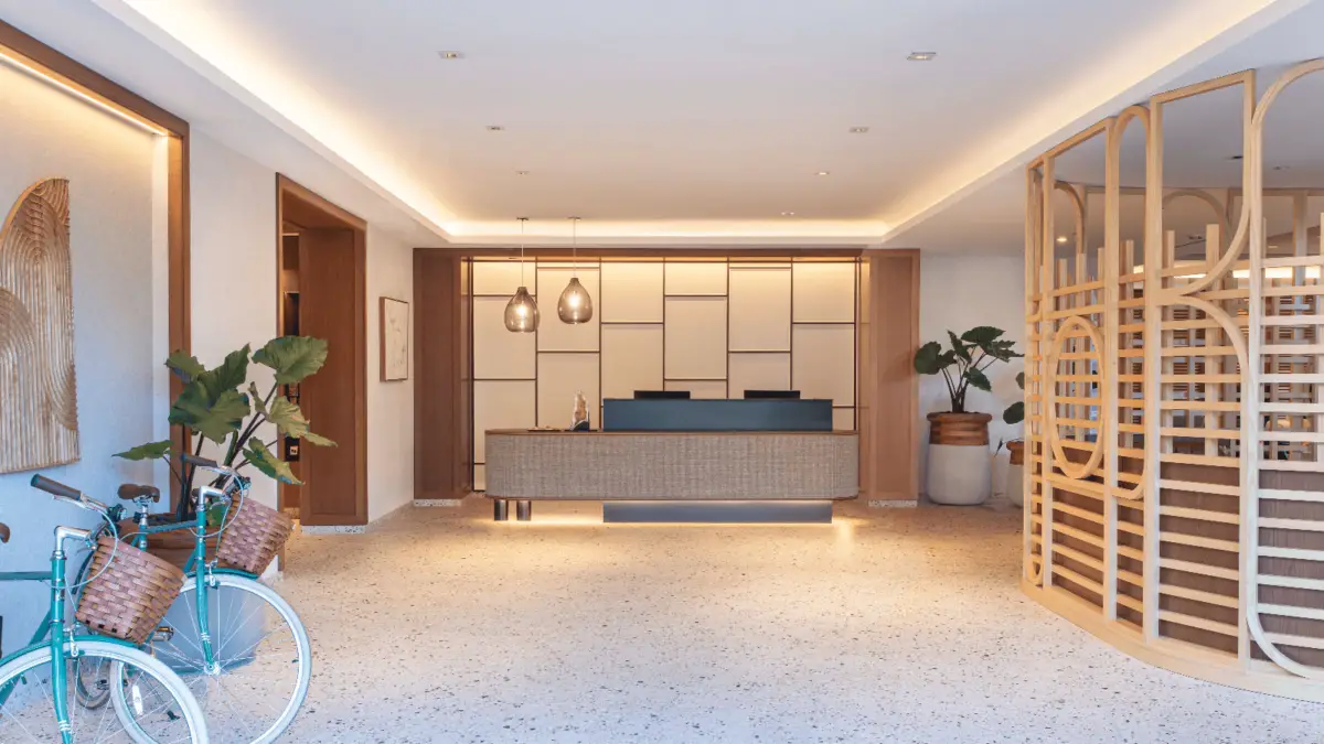 A modern hotel lobby with a minimalist design, featured among the Best Hip Hotels in the Bay Area. The central reception desk is illuminated by pendant lights. There are potted plants and a teal bicycle with a wicker basket on the left, while on the right stands a stylish wooden partition with geometric patterns.