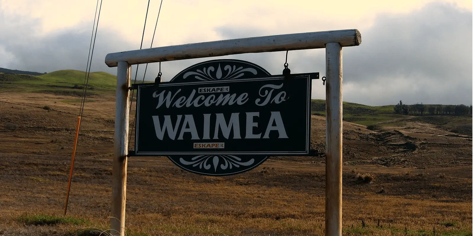 A wooden sign that reads "Welcome to Waimea" stands in a grassy field with hills in the background. The sky is cloudy, creating a serene and rustic atmosphere. Power lines are visible on the left side, and the sign includes the logo "ESCAPE," perfect for starting your food tour Waimea adventure.