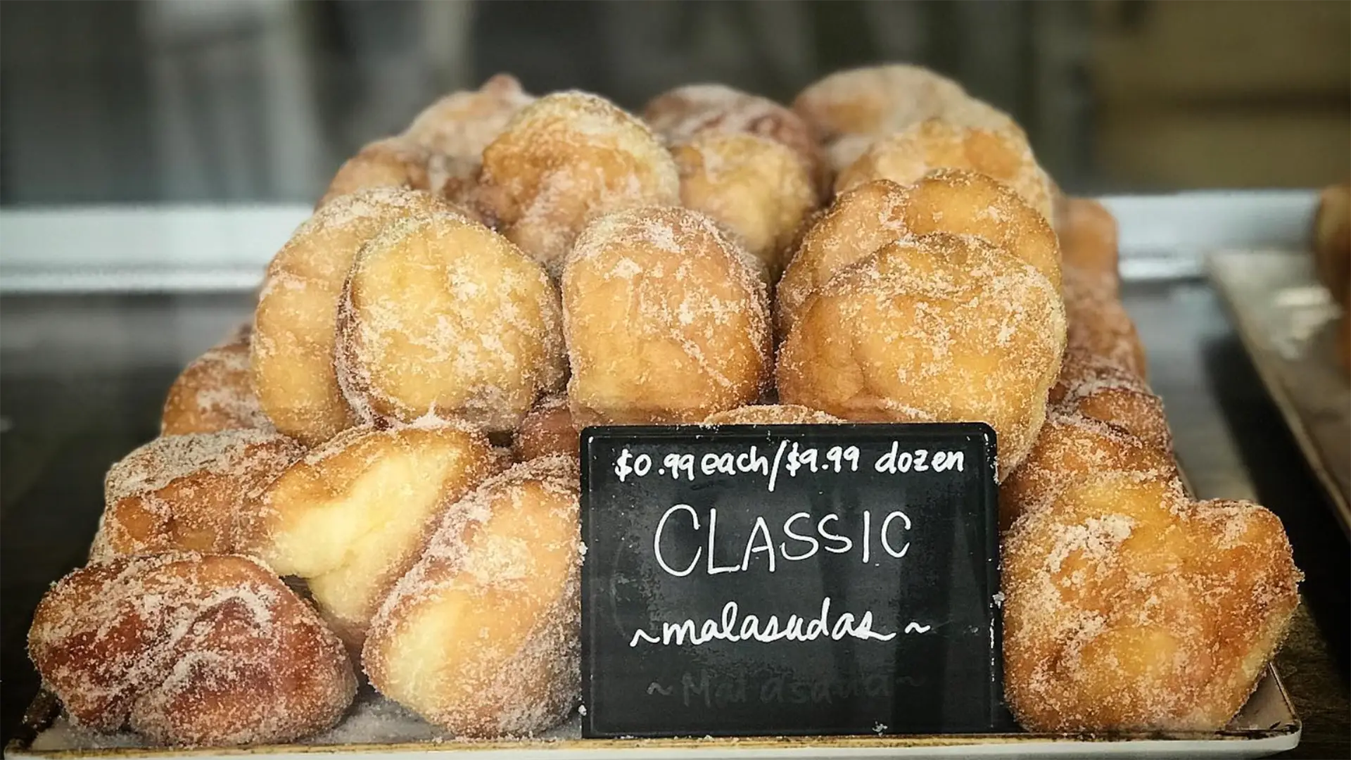 A display of sugar-coated malasadas (Portuguese-style fried doughnuts) is shown behind a glass case. A small chalkboard sign in front of them reads: "$0.99 each / $11.99 dozen - CLASSIC malasada." These are widely known as the best malasadas doughnut on Kauai, stacked in a pyramid shape.