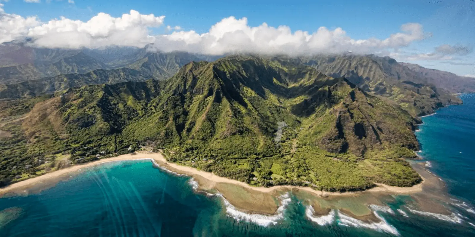 A scenic aerial view of a lush, green mountain range on the coast, with turquoise ocean waters washing up on sandy beaches. Rugged cliffs and dense vegetation are visible, while fluffy clouds hover over the peaks of the mountains in the background—truly the best island tour Oahu has to offer.