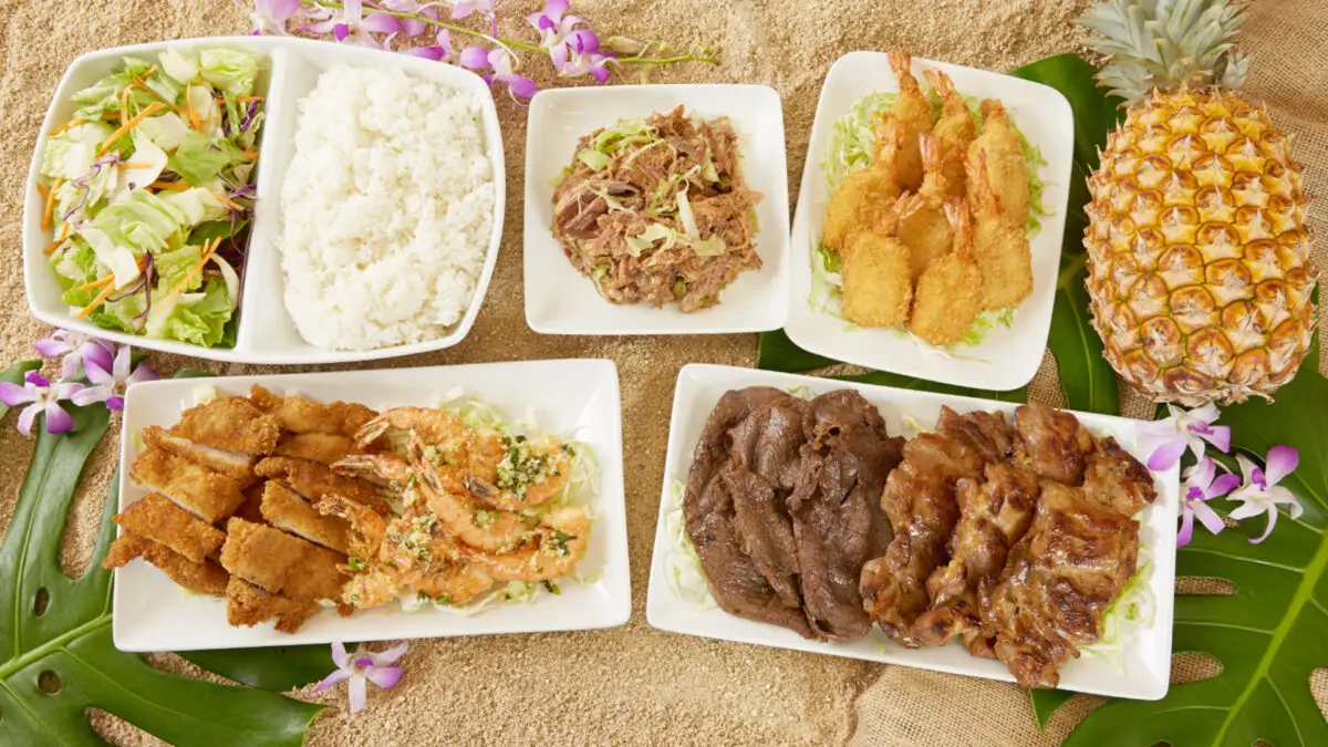 An assortment of dishes including white rice, shredded meat, fried shrimp, breaded cutlets, grilled meats, and vegetable salad served on rectangular white plates, arranged on a sandy surface with a pineapple and purple flowers as decoration. This spread showcases the best Hawaiian food Kauai has to offer.