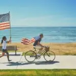 A person rides a bicycle along a seaside path, with an American flag waving from the back. Another person runs alongside, energetically holding a large American flag. The ocean and blue sky provide a scenic backdrop—one of the best things to do in the Bay Area Labor Day Weekend.