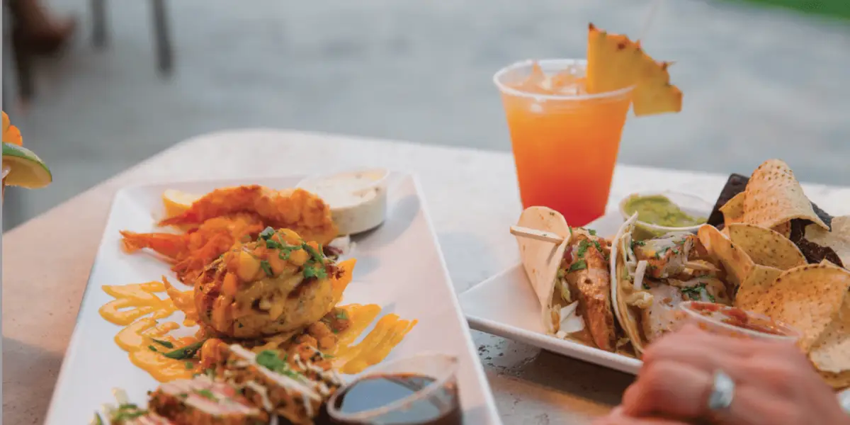 Two white plates of food and a drink on a table at the best lunch spot in Ko Olina, Oahu. One plate has assorted appetizers with sauces. The other plate has tacos with chips and guacamole. A fruity cocktail with an orange hue and pineapple garnish is next to the plates. A hand is reaching towards the food.