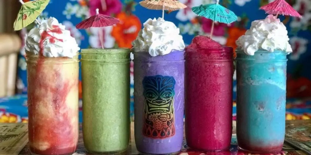 A row of five colorful tropical drinks in jars, topped with whipped cream and small paper umbrellas. The drinks are yellow, green, purple (with a Tiki face on the jar), pink, and blue, against a vibrant floral background—truly the best smoothies on Hawaii Island!