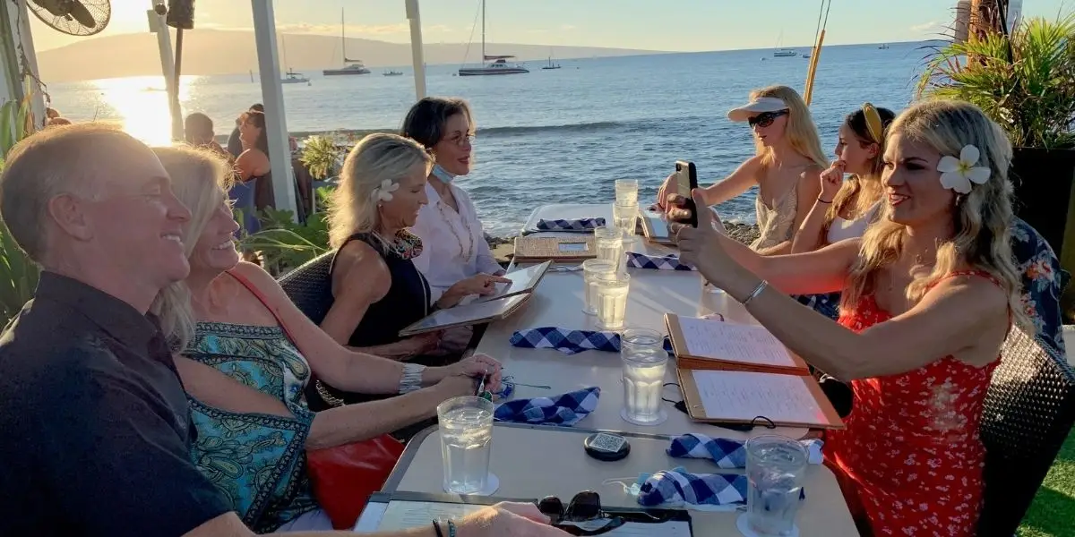 A group of six people sitting at an outdoor restaurant by the ocean, with the best sunset behind them. A woman in a red dress is taking a selfie with the group, who are all smiling and enjoying the moment. Sailboats can be seen in the background on the water, encapsulating perfect Sunset Dining in Maui.