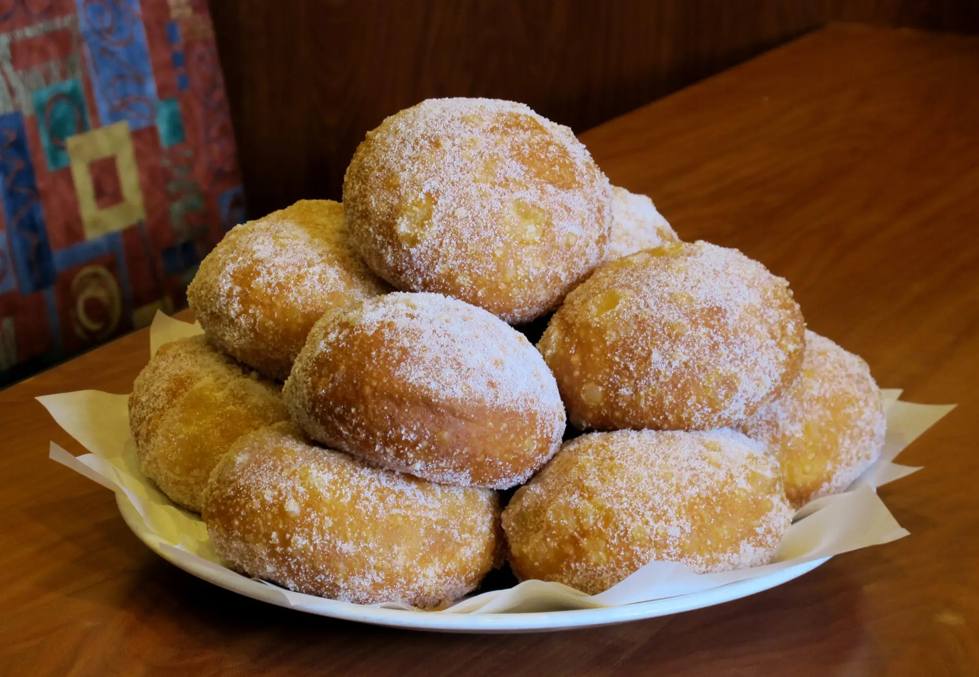 A white plate is piled high with the best malasadas on the Big Island, sitting on a wooden table. The golden-brown pastries are generously covered with granulated sugar, creating a tempting and delicious appearance.