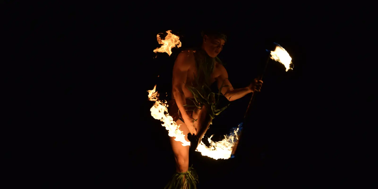 A performer in traditional attire spins a flaming torch, creating a captivating fire dance against a dark background. Flames encircle the performer's body, illuminating the figure and emphasizing the dynamic movement and intensity of one of the top luaus Maui has to offer.