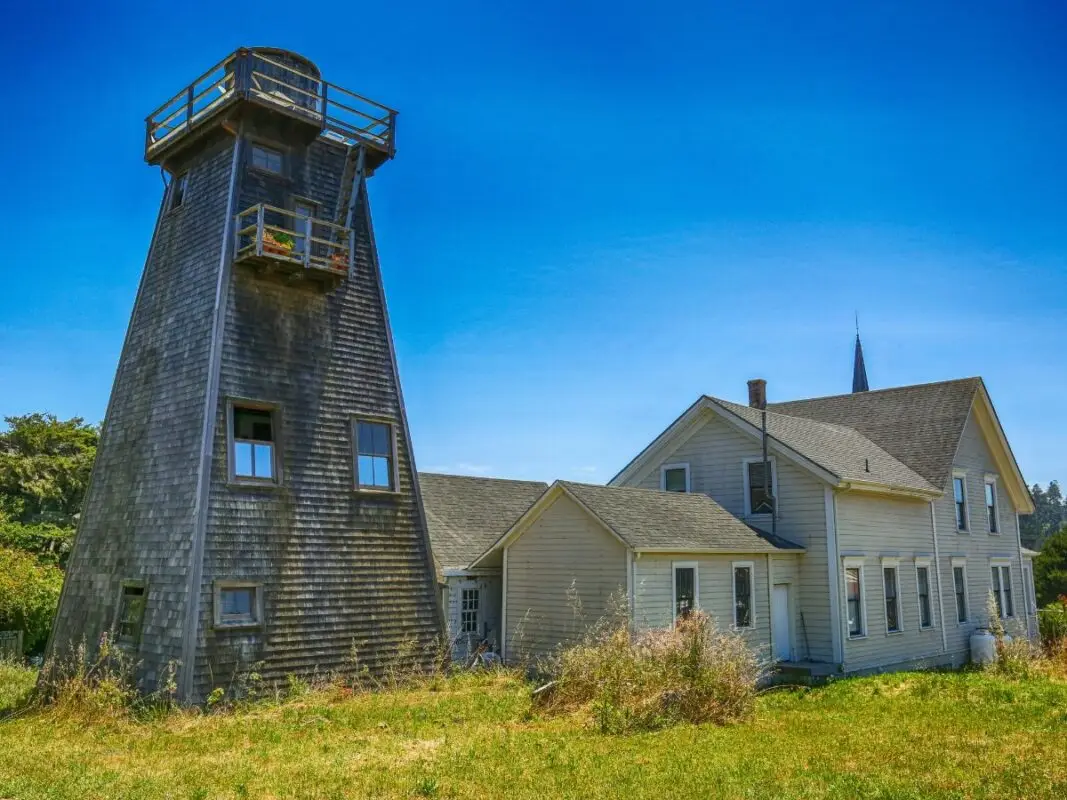 A weathered, wooden lighthouse tower with an elevated observation deck stands next to an old, two-story house on the Mendocino Coast. The structures are surrounded by overgrown grass, and the sky is clear, vibrant blue. In the background, a tall spire is visible.