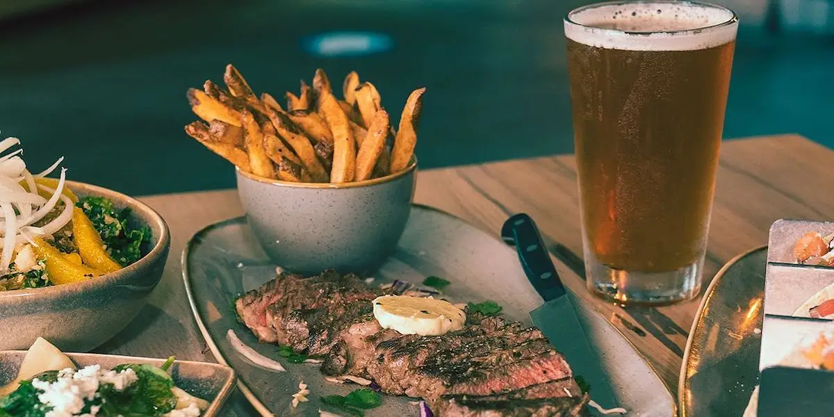 A wooden table displays a meal with a serving of steak topped with a round of butter, a side of crispy fries in a bowl, a fresh mixed salad, and a glass of beer. It's the best happy hour Oahu has to offer. A knife rests next to the steak on the plate, while the background is softly blurred.