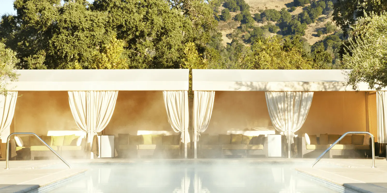 A luxurious outdoor pool area with mist rising from the water. In the background are cabanas draped with light fabric, providing shade and seating with cushions. Beyond the cabanas, green trees and a mountainous landscape complete the serene setting at one of Napa's Best Spa Wellness Hotels.