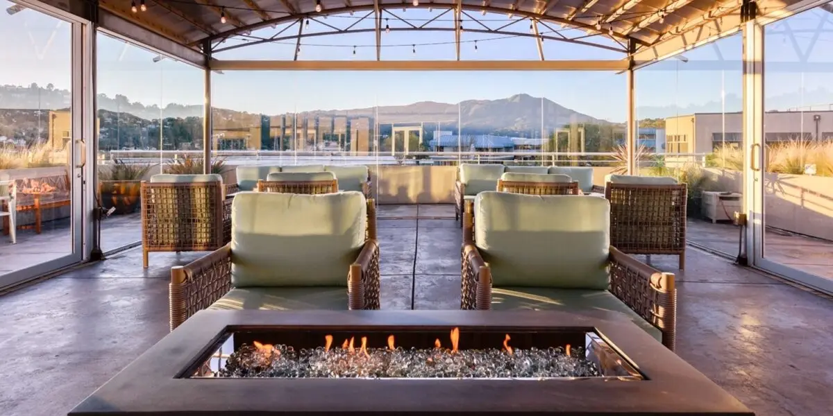 A rooftop patio with wicker chairs and beige cushions arranged around a rectangular glass fire pit. The area is enclosed by glass walls, offering views of distant mountains and blue skies. This inviting space, often found in the best value hotels North Bay, boasts a relaxed ambiance with natural lighting.