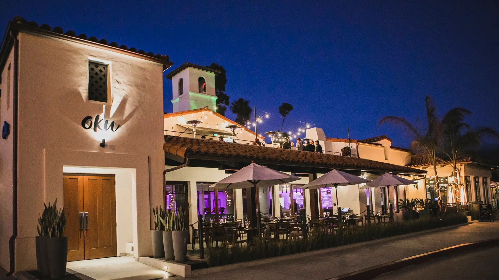 A charming restaurant illuminated at night, featuring outdoor seating with white umbrellas. The building has a Mediterranean architectural style with a central tower adorned with string lights. Known as one of the best dinner places in Santa Barbara, the sign "oku" is displayed above the main entrance.