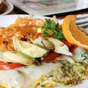 A plate of breakfast food reminiscent of Monterey Peninsula restaurants features a veggie omelet with melted cheese, topped with avocado slices, tomatoes, and a sprig of parsley. The dish is accompanied by crispy hash browns and garnished with an orange slice. A fork is visible to the side.